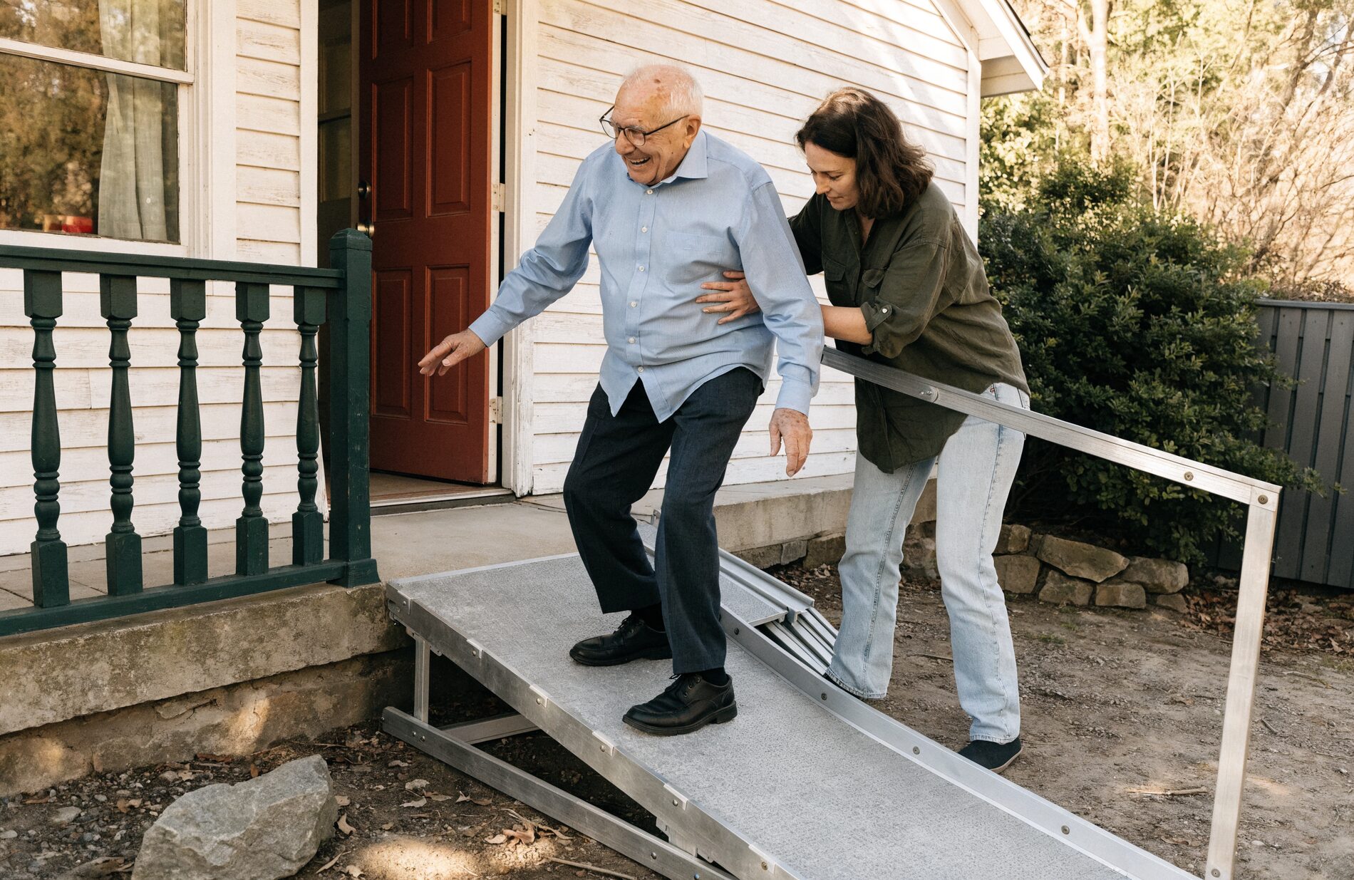 A daughter steadies her father as he walks a wooden ramp into the house at late afternoon light